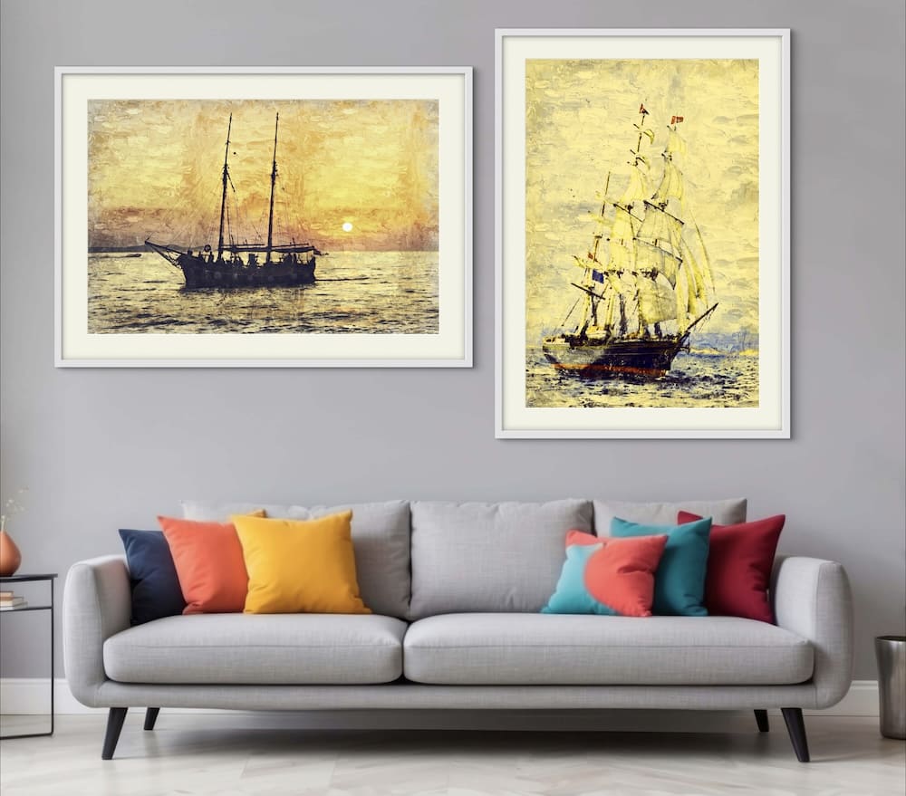 Two framed oil paintings of sailing ships hanging on a grey wall above a double seater lounge chair.