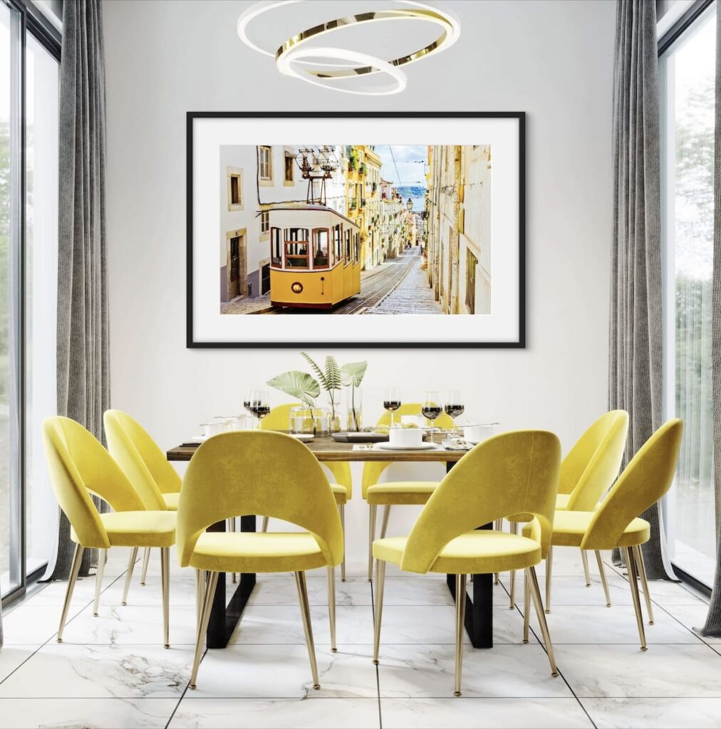 Framed photo a yellow tram making its way down a street. The street is lined with buildings. The picture is hanging on a wall above a dinning table with lime chairs