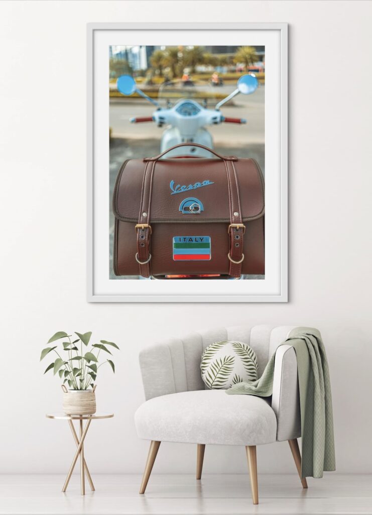 Framed image of a Vespa  with a leather carry bag hanging on a wall above a lounge chair.