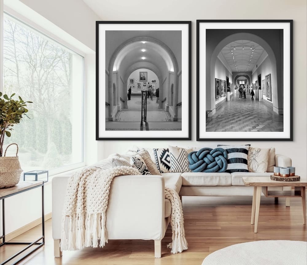 The image shows a modern living room with a large window on the left side, allowing natural light to flood the space. The room features a white sofa adorned with various patterned cushions and a knitted throw blanket. Above the sofa, there are two large black-and-white framed photographs of what appears to be an art gallery or museum. The left photograph shows a staircase leading up to a gallery with framed artworks on the walls, while the right photograph depicts a long hallway lined with paintings and people walking through it. The room also includes a wooden side table with a plant and a woven basket, adding a touch of nature to the decor. The overall aesthetic is clean, modern, and inviting.
