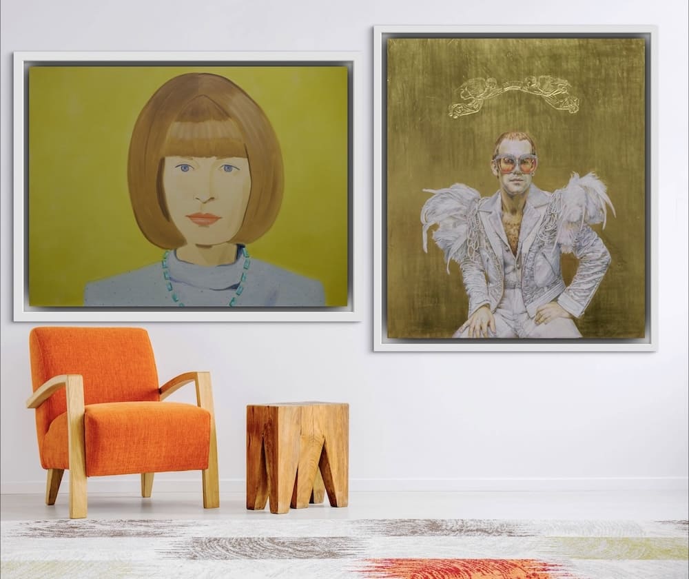 A modern interior featuring two framed portraits on the wall. The left artwork depicts a stylized painting of a woman with a bob hairstyle, wearing a light top and a turquoise necklace against a vibrant yellow background. The right artwork portrays a flamboyant man in a white feathered suit with oversized glasses, set against a golden background with intricate detailing above him. Below the artworks, a cozy orange armchair and a sculptural wooden side table add warmth and texture to the space.