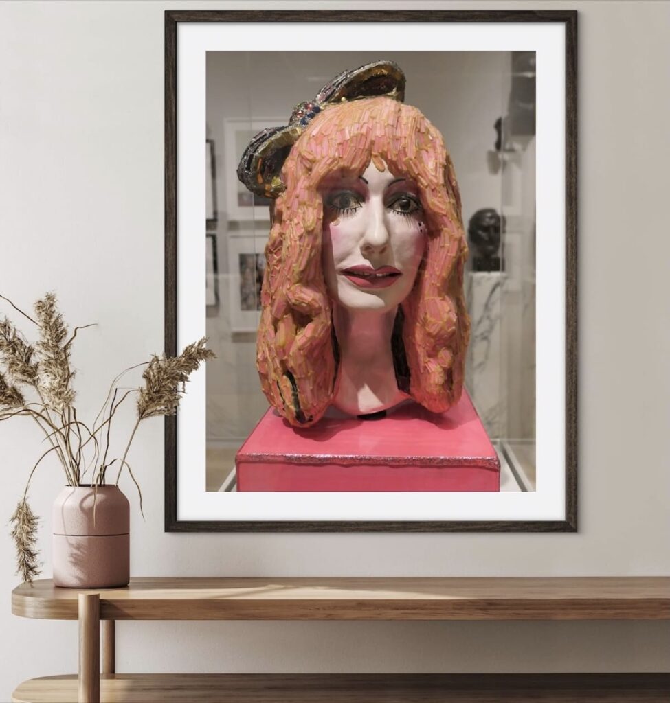 A framed photograph of a sculpture is displayed on a wall above a wooden shelf with a vase of dried plants. The sculpture features a head with exaggerated, textured hair and a hat, all in vibrant colors. The background shows a gallery setting with other artworks