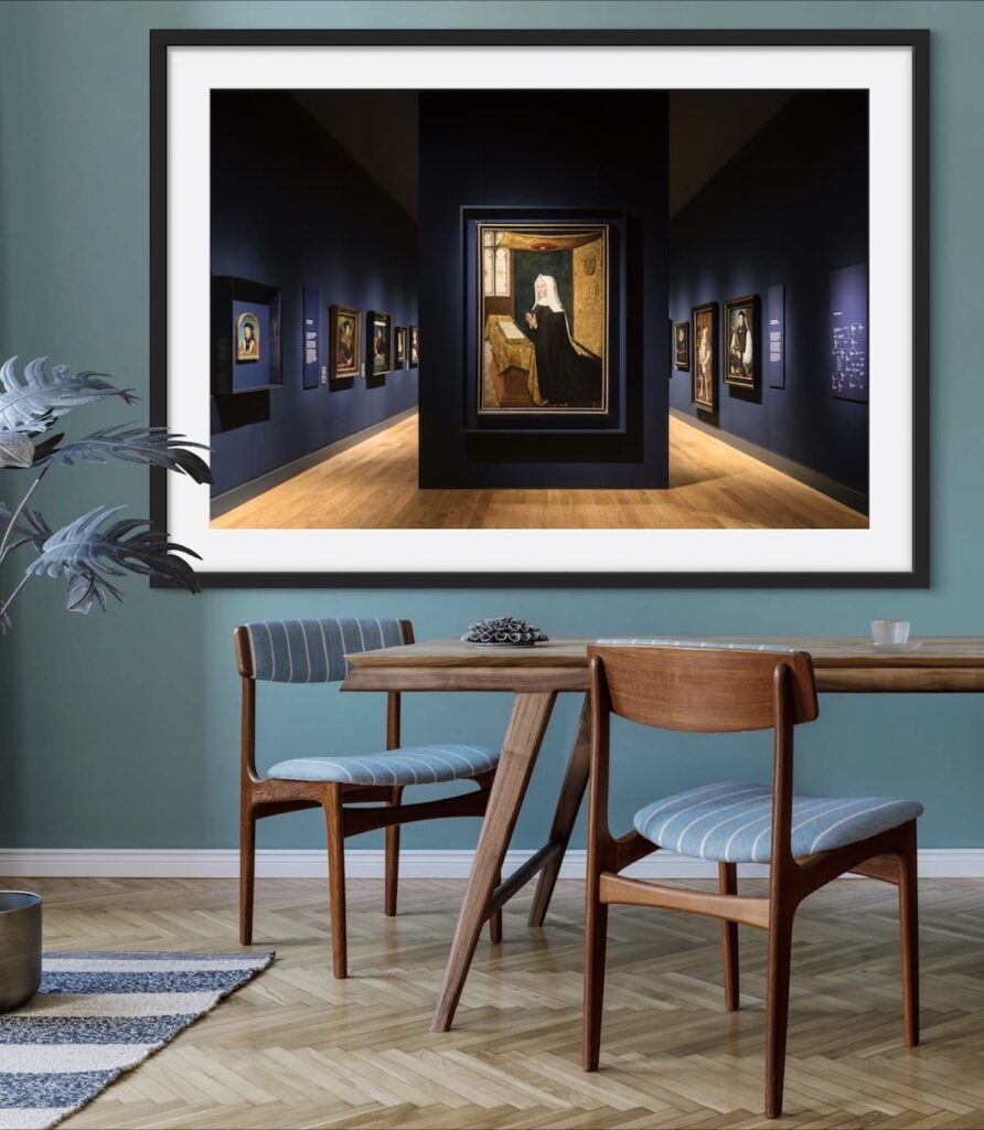 A stylish dining area featuring a framed photograph of a dark blue gallery room showcasing classic paintings, prominently displaying a central artwork of a veiled figure. The interior is complemented by mid-century modern furniture, including a wooden dining table and chairs upholstered in light blue striped fabric. A green accent wall and natural wood flooring complete the inviting and sophisticated atmosphere, with a potted plant adding a touch of greenery.