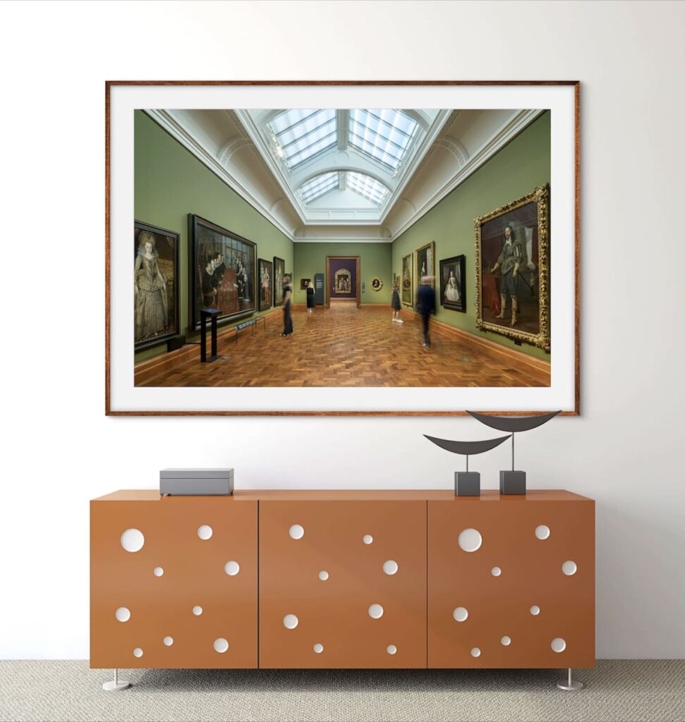 An elegant gallery setting with a large framed photograph of an art museum's interior, featuring a green-walled exhibition room filled with paintings, displayed above a modern orange sideboard adorned with circular cutouts. The sideboard is accessorized with minimalistic sculptures, creating a sophisticated and artistic ambiance.
