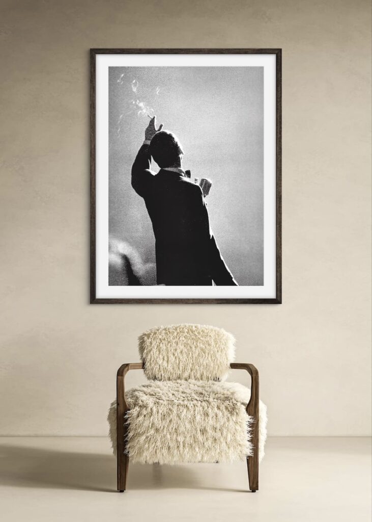 Black and white photo taken from the back of Frank Sinatra at a singing mic with a raised hand holding a cigarette 