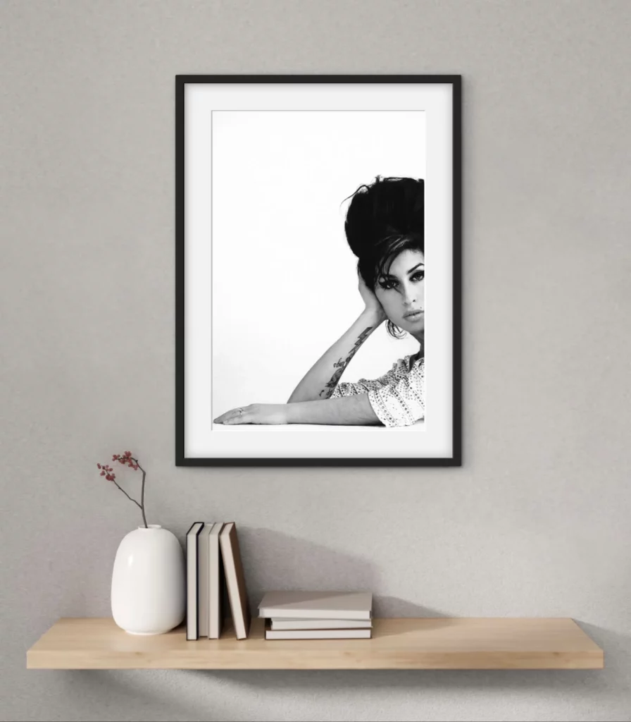 Minimal interior with a floating wooden shelf, books, and a white ceramic vase beneath a framed black-and-white portrait of a woman with a beehive hairstyle, resting her head on one hand and gazing toward the camera against a clean white background.