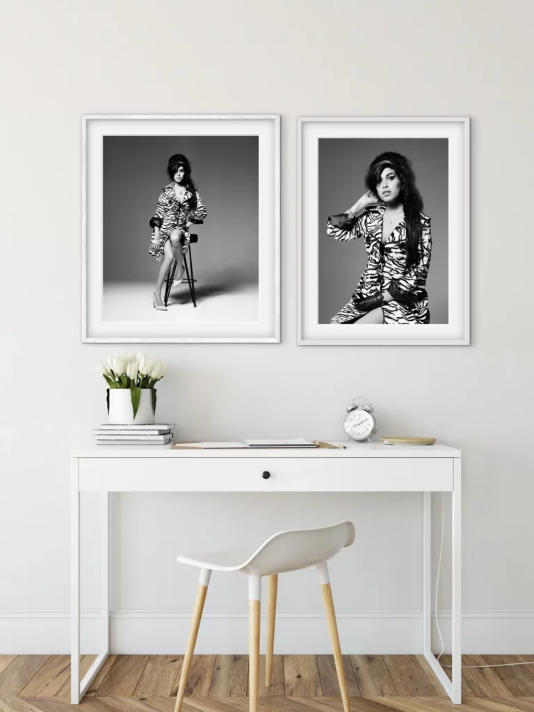 Bright, minimalist home office with a white desk, chair, flowers, and a clock, featuring two framed black-and-white portraits of a woman in a patterned dress. One portrait shows her seated on a stool, while the other is a closer portrait with her hand resting behind her neck.