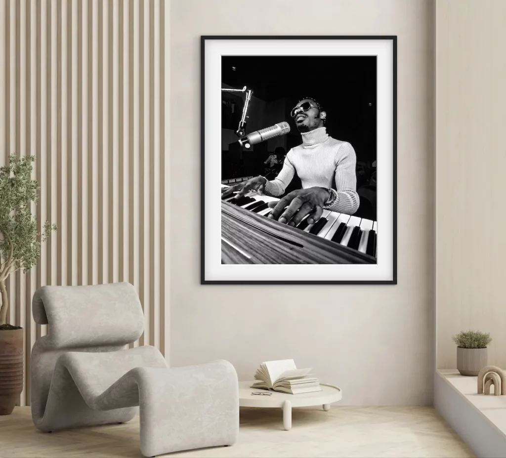 Modern, neutral-toned reading nook with a sculptural lounge chair and small side table, featuring a large framed black-and-white photograph of a man wearing sunglasses, singing into a microphone while playing a keyboard.