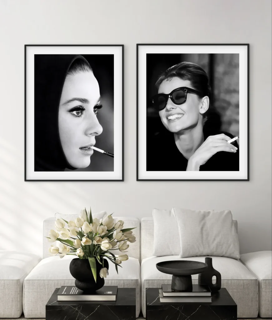 Two framed black-and-white portraits of Audrey Hepburn seen smoking hanging on a wall above a white sofa, with a black vase of white tulips resting on a coffee table in the foreground.