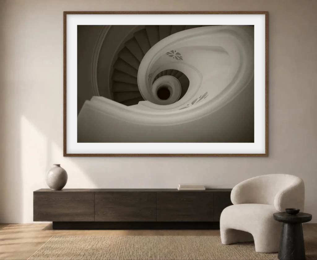 A framed black-and-white photograph of a spiral staircase viewed from above, displayed on a dark wooden console table next to a modern, cream-colored boucle armchair and a round black side table.