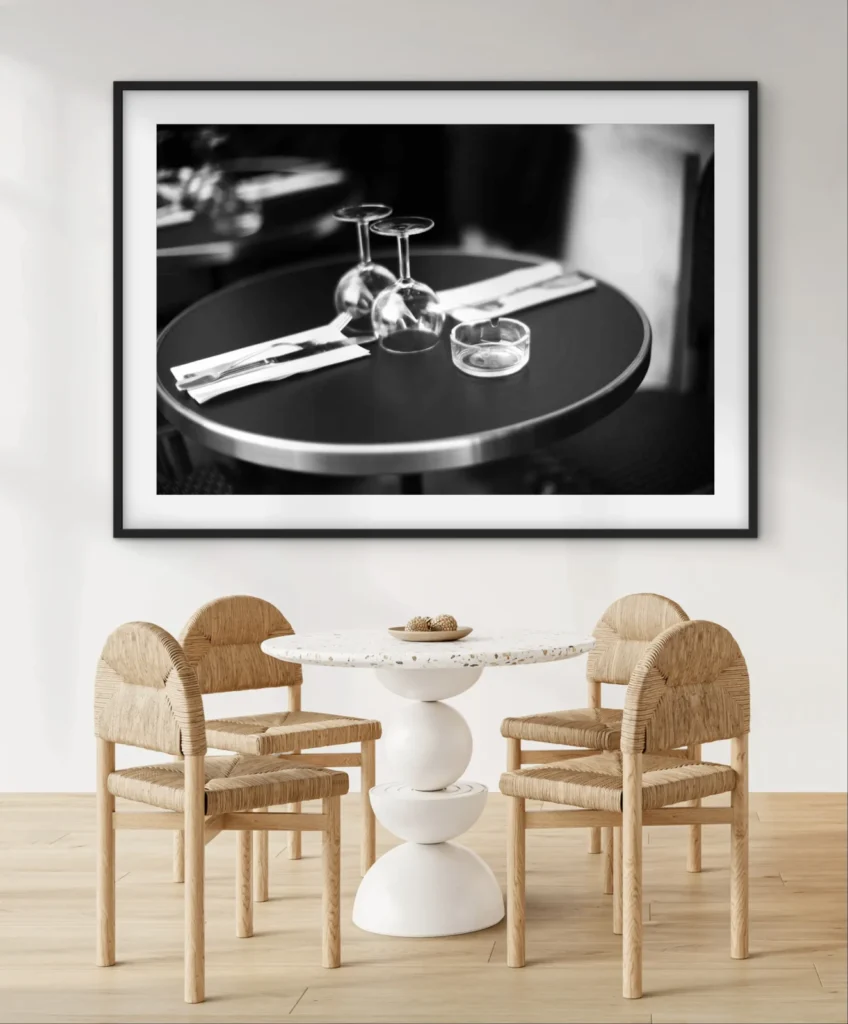 A framed black-and-white photograph of a round table set with two wine glasses, a small bowl, and folded napkins.