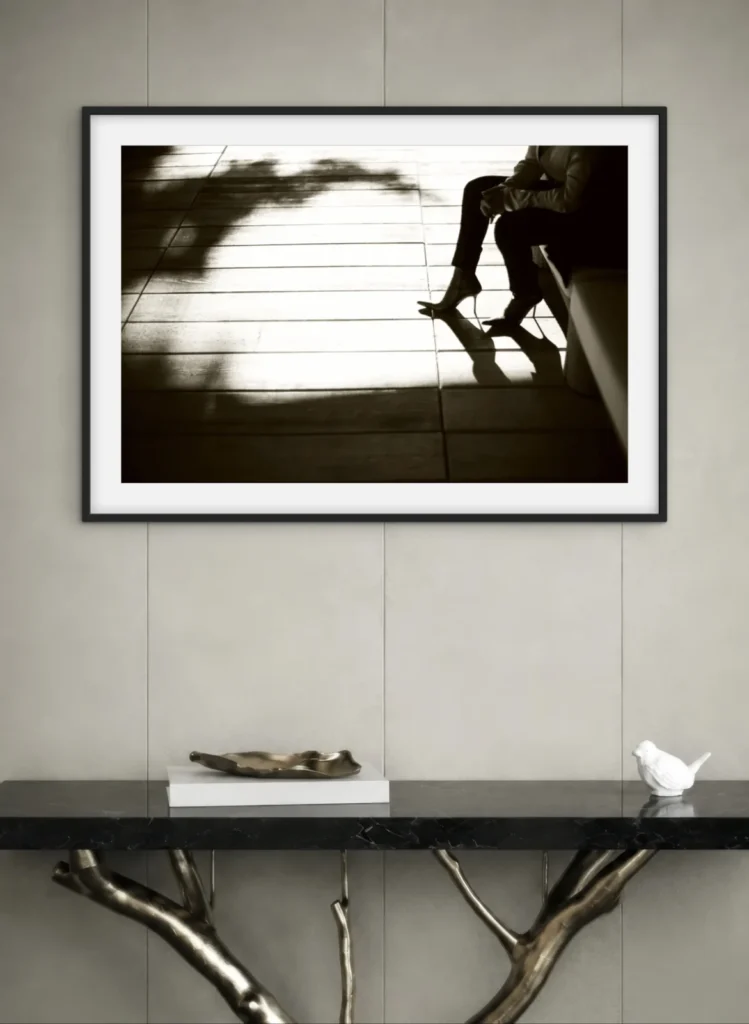 This image features a framed black and white photograph hanging on a light-colored wall. The photograph depicts a person sitting near a window, with strong shadows cast across the floor. Below the frame, there is a dark console table with a distinctive, branch-like metallic base, displaying a small white bird figurine and a decorative bowl on top.