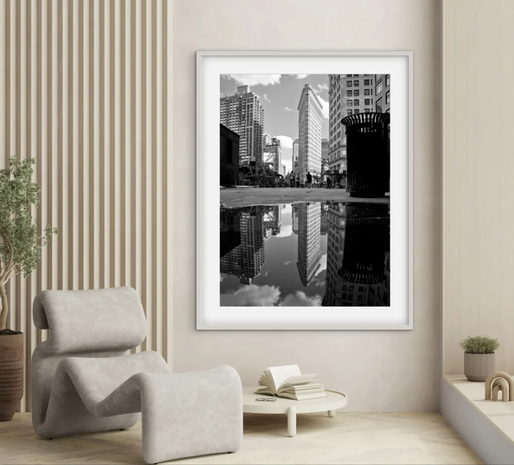 This image features a framed black and white photograph of a city street scene, with a trash can in the foreground and tall buildings reflected in a puddle on the street. The photograph is displayed in a modern living room setting, next to a uniquely shaped, plush beige chair and a small round coffee table with an open book on it.