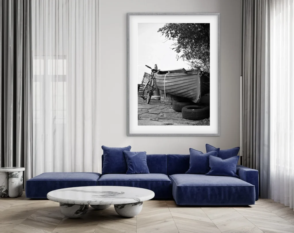 This image shows a framed black and white photograph of a boat and a bicycle, hanging on a wall above a blue velvet sofa and a marble coffee table.