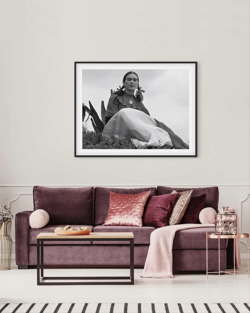 A black-and-white framed portrait of Frida Kahlo sitting outdoors among plants, displayed above a plush purple sofa.