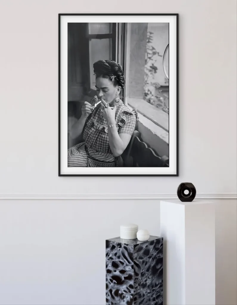 A black-and-white framed portrait of Frida Kahlo sitting by a window smoking a cigarette, displayed above a black and white marble pedestal.