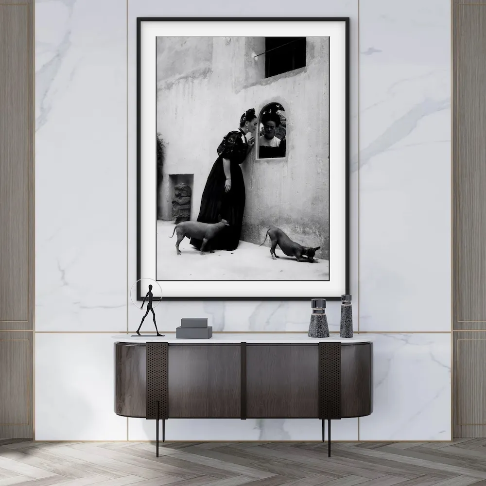 A black-and-white framed photograph of Frida Kahlo standing outside with two dogs, displayed above a modern dark wood console table.