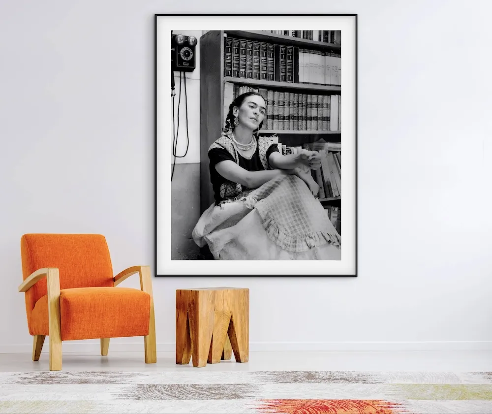 A black-and-white framed portrait of Frida Kahlo sitting in front of a bookshelf, displayed above an orange armchair and a wooden side table.