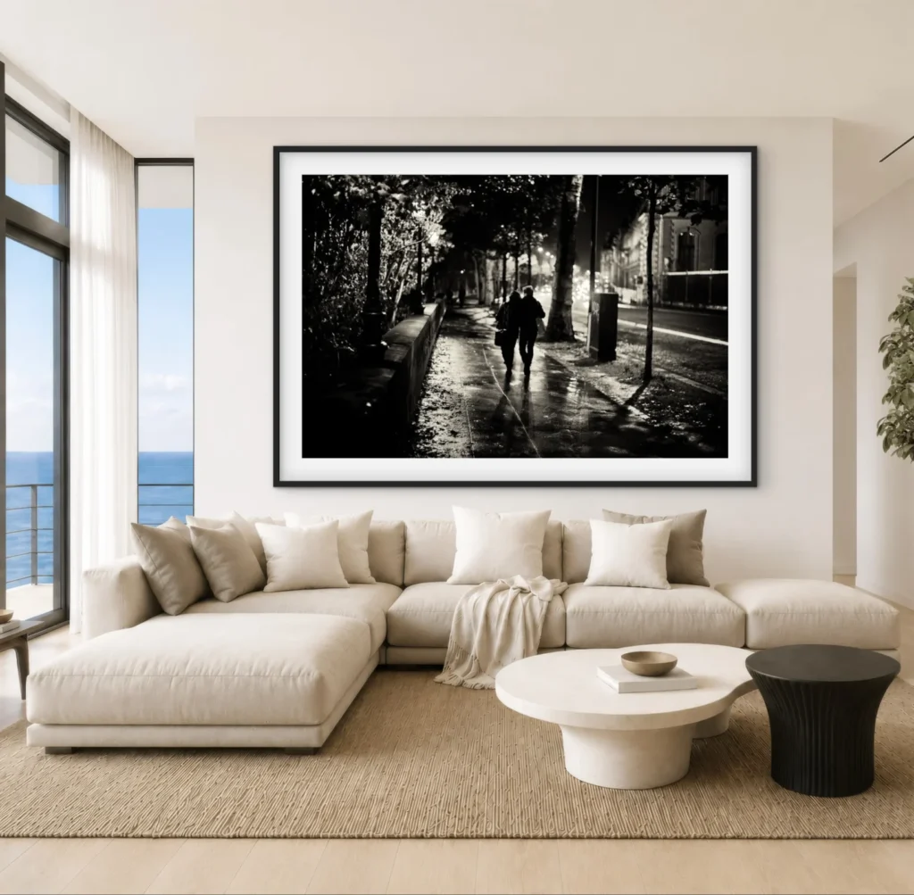 This image shows a modern living room featuring a large, beige L-shaped sectional sofa facing a black-and-white framed photograph of a couple walking down a tree-lined path. The room also includes a round, two-tiered coffee table and a large window with a view of the ocean.