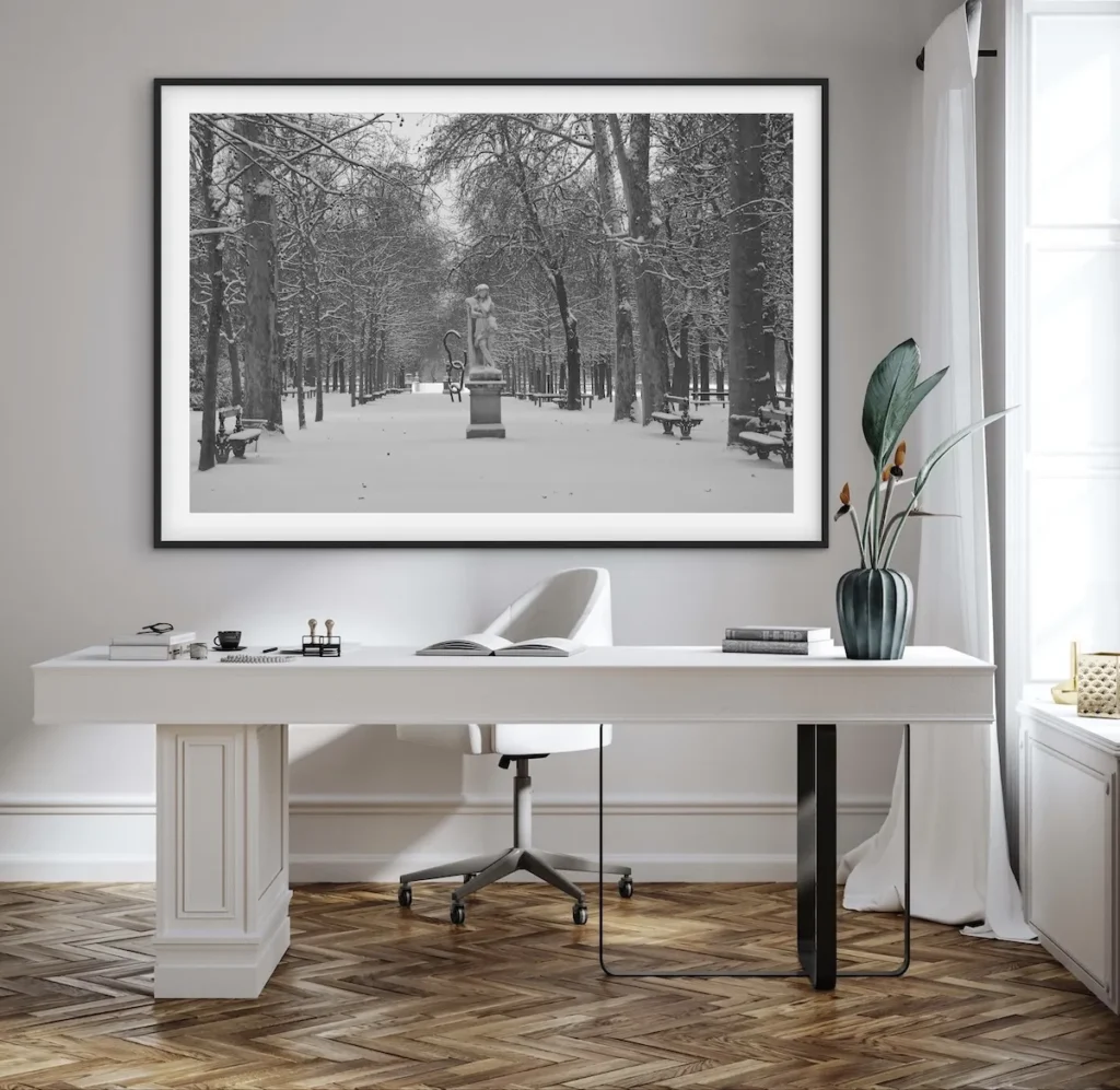 A framed black-and-white photograph of a snow-covered park path lined with bare trees and a central statue, displayed above a modern white desk.