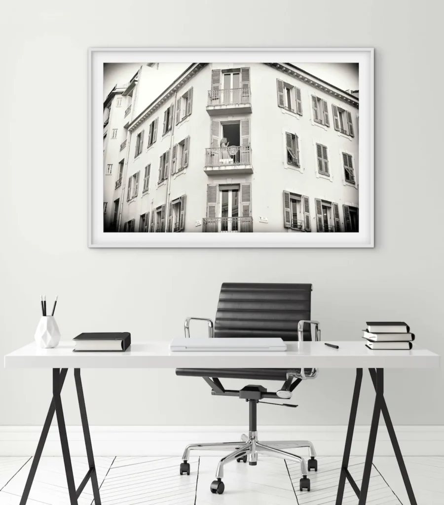A framed black-and-white photograph of a multi-story building with shuttered windows and small balconies, displayed above a white desk with a black office chair.