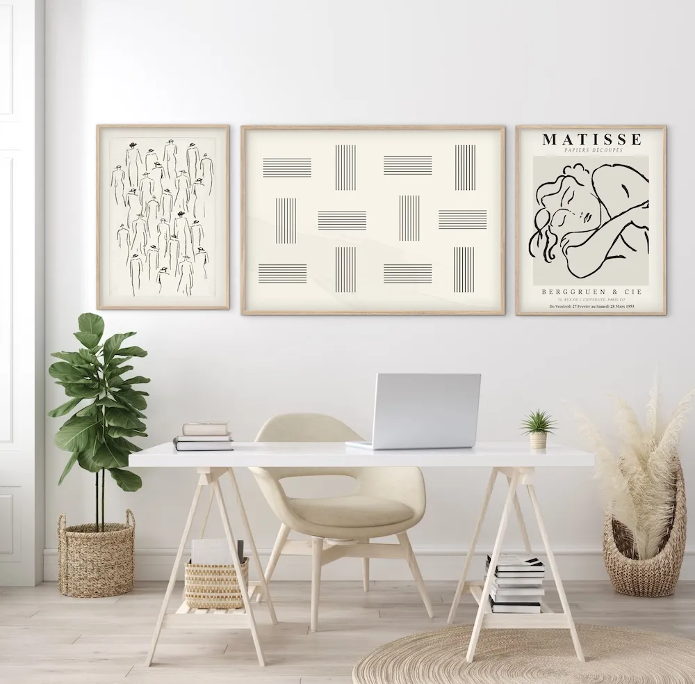 Three framed art prints—one featuring abstract figures, one with a geometric line pattern, and a Matisse exhibition poster—displayed above a white trestle desk with a laptop.