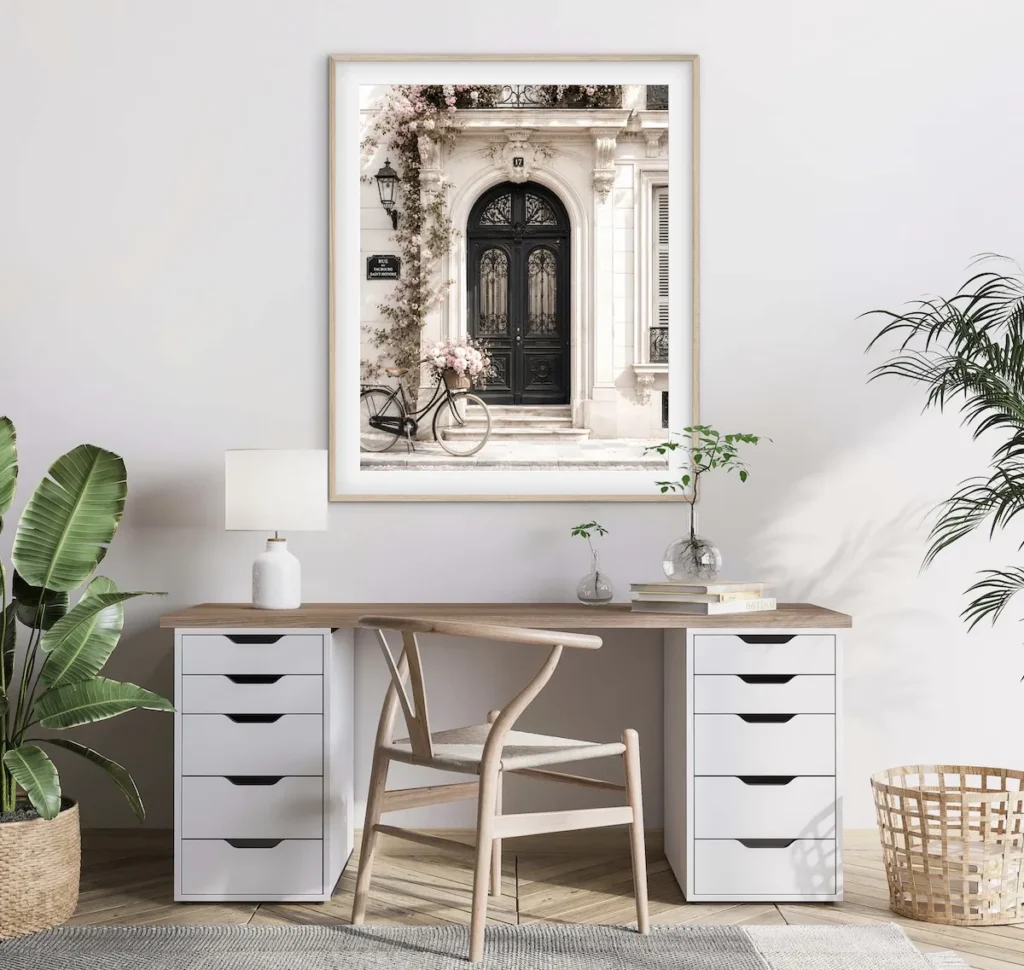 A framed photograph of a black bicycle with a flower basket parked in front of an ornate black double door, displayed above a white desk with two drawer units and a wooden chair.
